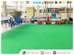 ลานกิจกรรมกลางแจ้งในร่ม ปูพรมสีเขียวสดเต็มพื้นที่ มีแถวเก้าอี้พลาสติกสีน้ำเงิน โต๊ะวางขวดน้ำดื่ม และอุปกรณ์เชียร์กีฬาหลากสีสัน
